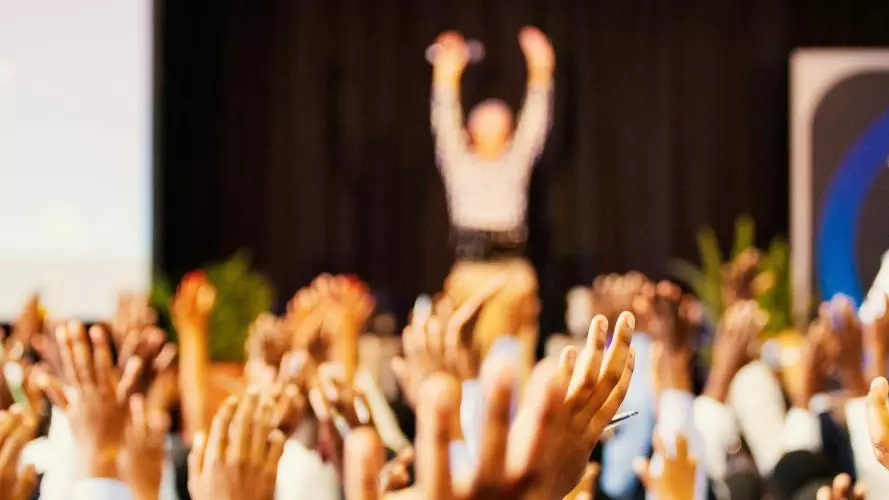 Blurry picture of audience raising hands in front of a stage where a speker stands. The photos is taken from the perspective of the audience.