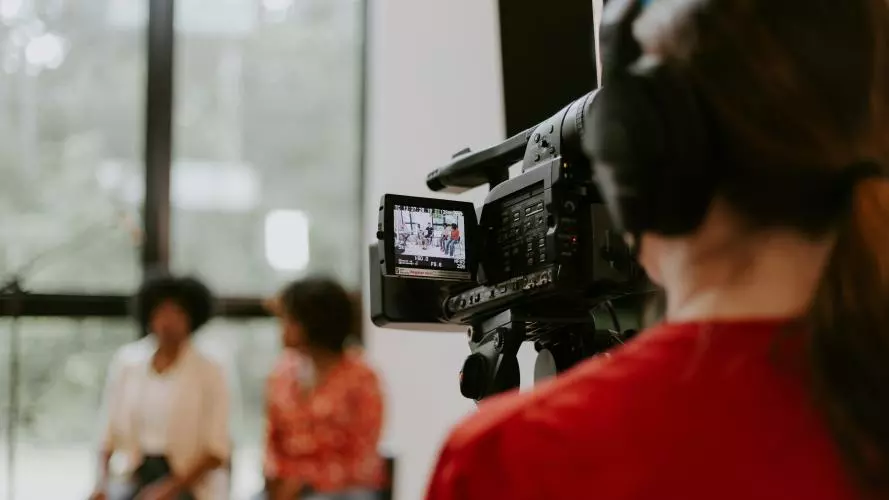 Man in red shirt holding black videocamera shooting two women in conversation.