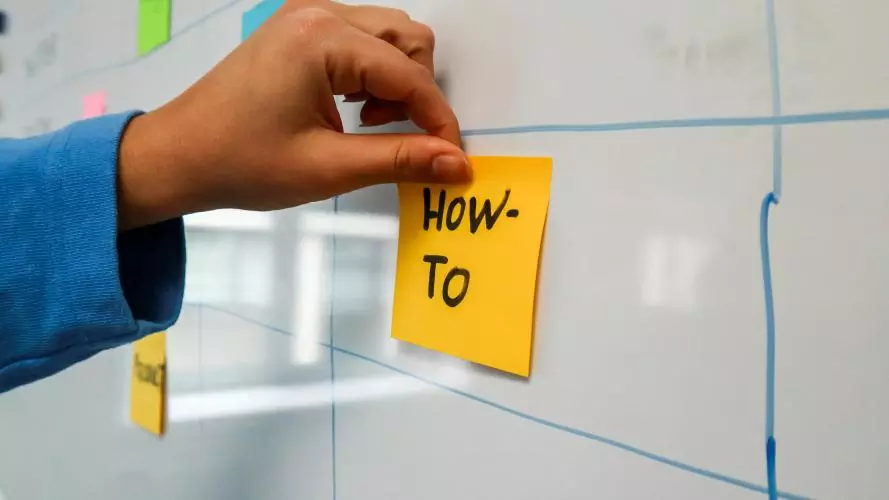 A hand sticking a note on a whiteboard with the words 'how to' written on it.
