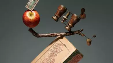 Absurd picture where a series of objects, including an apple, a pencil, a book, a pair of binoculars etc are impossibly balanced on top each other to prompt the viewer's interest.