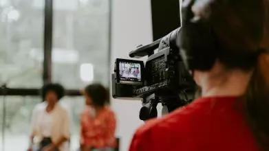 Man in red shirt holding black videocamera shooting two women in conversation.