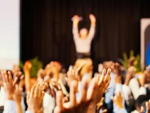 Blurry picture of audience raising hands in front of a stage where a speker stands. The photos is taken from the perspective of the audience.