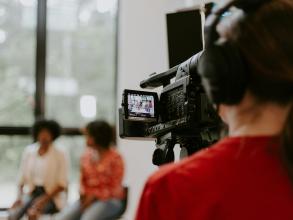 Man in red shirt holding black videocamera shooting two women in conversation.