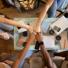 Four people bringing their fists together over a table with notepads and takeaway coffee cups